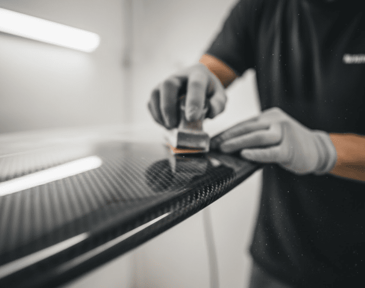 The technician uses fine sandpaper or resin tools to polish the edges of the surfboard, focusing on the hands and craftsmanship details. The soft light emphasizes the spirit of craftsmanship